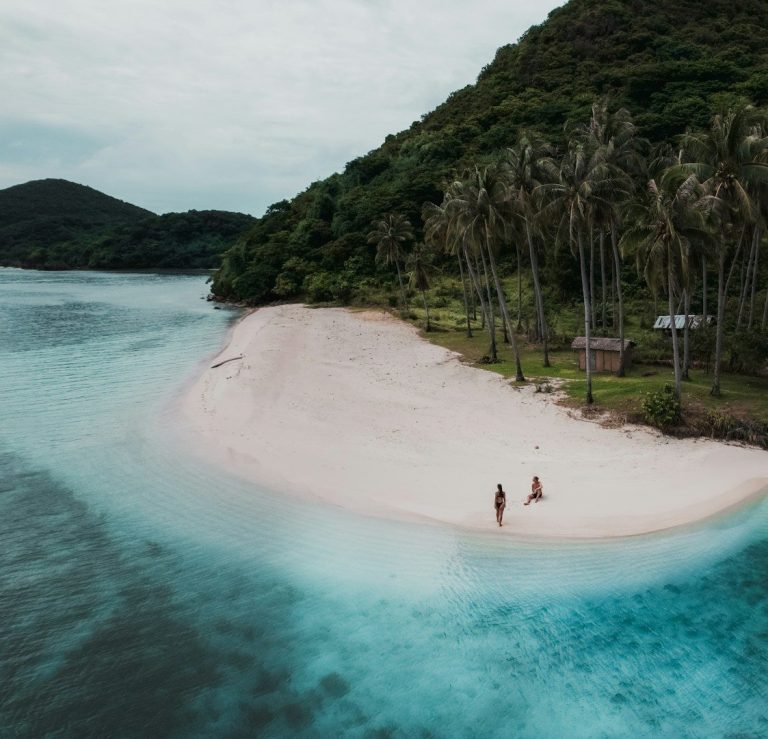 Plage de sable blanc, entourée d'eaux turquoises et de palmiers, avec deux personnes marchant.