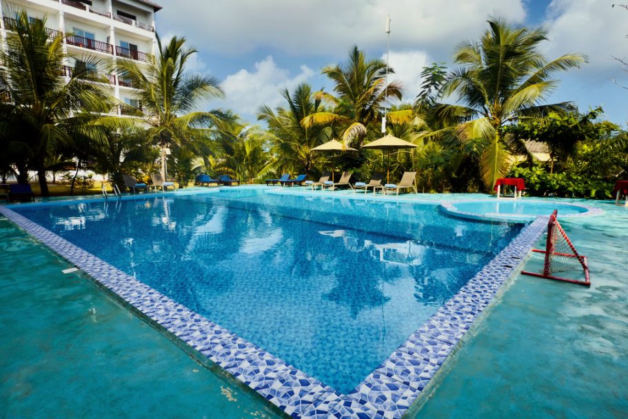 Piscine bleue entourée de palmiers et de chaises longues sous un ciel nuageux.