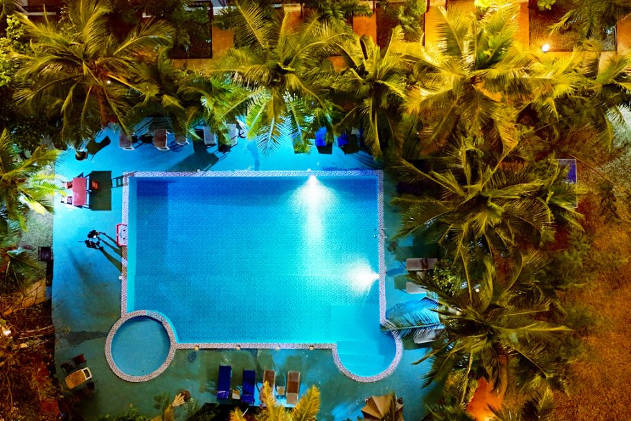 Piscine illuminée entourée de palmiers, avec des chaises longues autour.