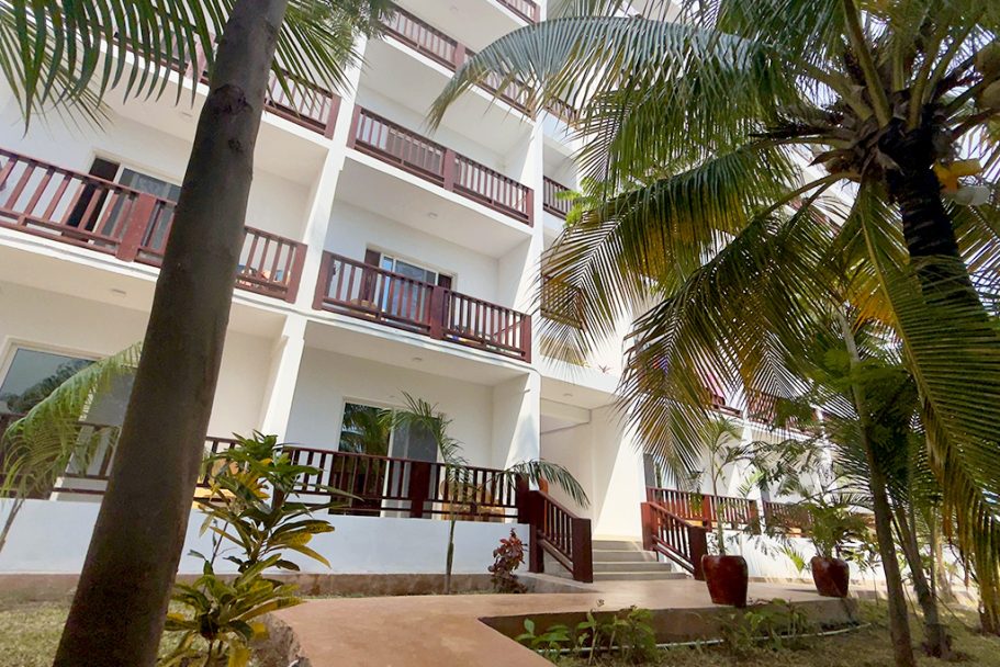 Bâtiment blanc à plusieurs étages avec balcons, entouré de palmiers verdoyants.