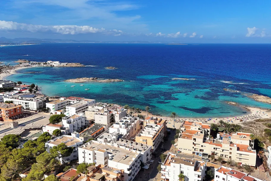 Vue aérienne d'une côte ensoleillée avec des eaux turquoise et des bâtiments en bord de mer.