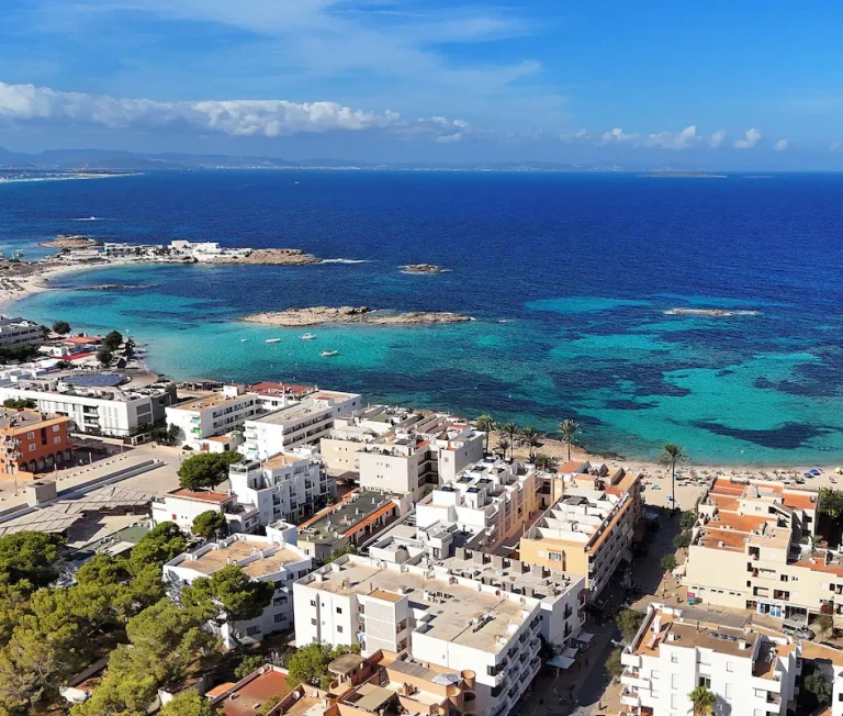 Vue panoramique d'une plage et de la mer turquoise, avec des bâtiments en arrière-plan.